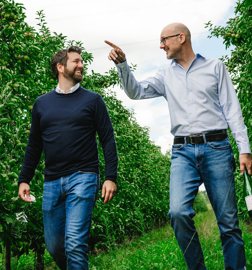 De twee makers van Klokhuys Fermenten wandelen lachend tussen de rijen fruitbomen.