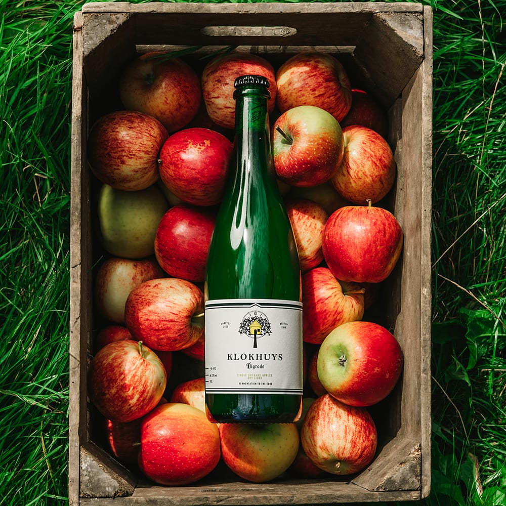 Een houten kist vol rode appels in het gras, met een fles Klokhuys in het midden — van bovenaf gefotografeerd.